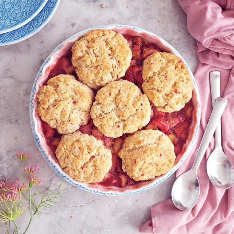 functional image strawberry rhubarb cobbler top down uncut with blue plates, pink flowers and pink fabric around it.