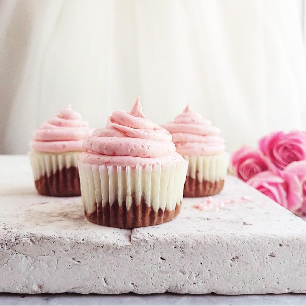 functional image 3 neapolitan cupcakes on a white marble block side view with pink roses in the background you can see the vanilla and chocolate cupcake through the white cupcake liner strawberry frosting on top