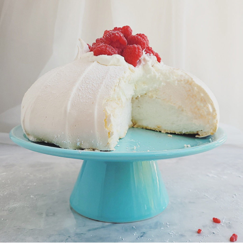 functional image pavlova with berries on a blue cake stand with one slice missing so you can see the interior layers of meringue and whipped cream