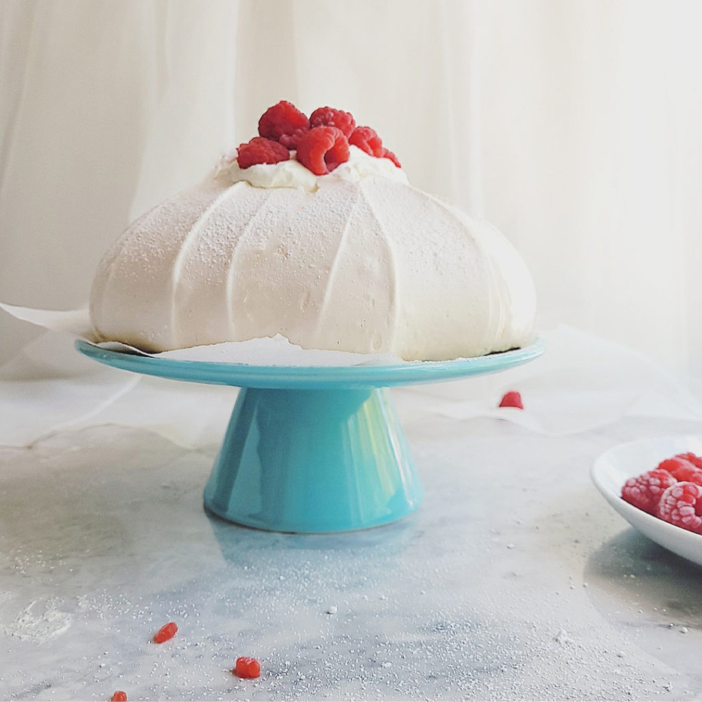 pavlova main image side view on a teal blue cake stand with whipped cream and red raspberries on top
