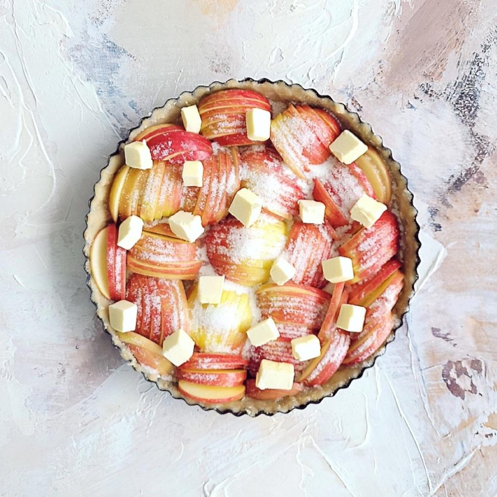rustic apple tart unbaked with sugar and cubed butter top down on a painted canvas