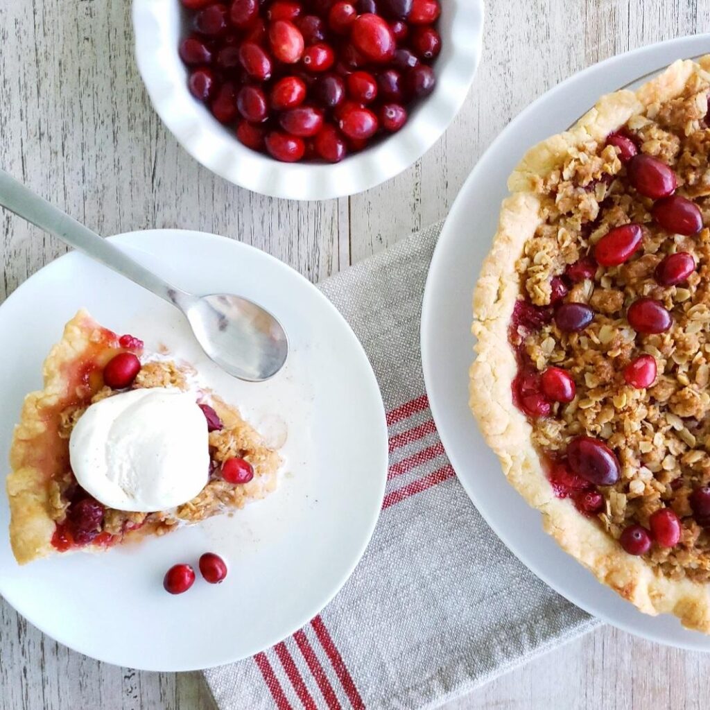 functional image top down on white washed wood. cranberry apple crumble pie slice of pie on a place with ice cream a bowl of fresh cranberries and a half moon view of the pie