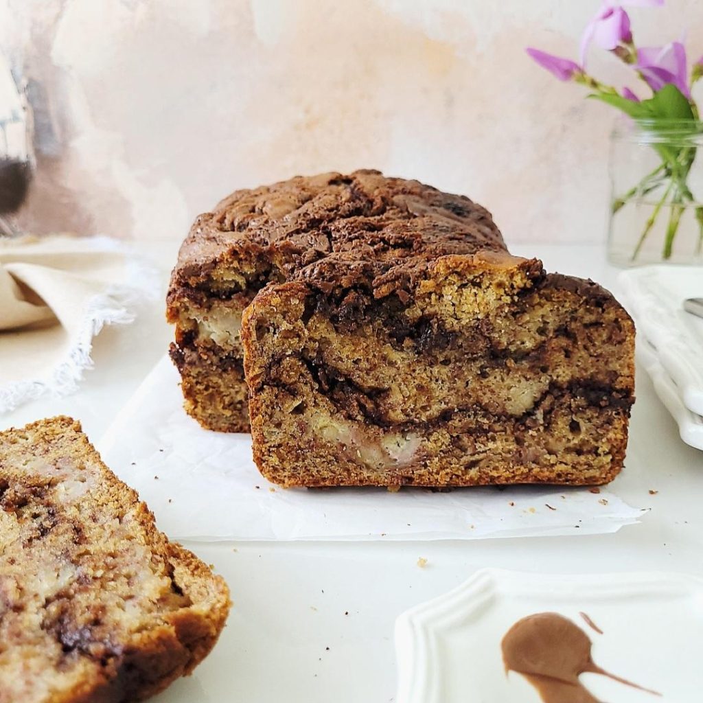 functional image nutella banana bread side view of loaf with one slice askew and other slice in the corner. styled with fresh cut purple flowers in a glass vase, white plates background is peach abstract 