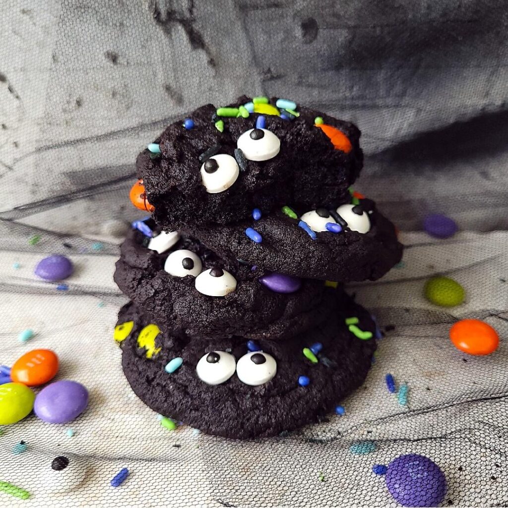 functional image m&m halloween cookies with candy eyes and halloween sprinkles. 4 cookies stacked on top of each other on black tulle with a distressed gray background