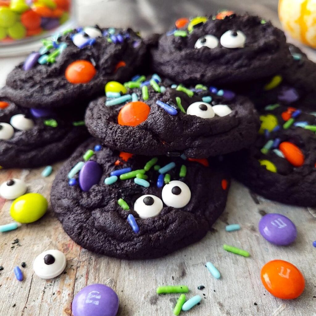 functional image chocolate halloween cookies side view of black cookies piled on top of each other cookies are garnished with candy eyes, halloween m&ms and halloween sprinkles