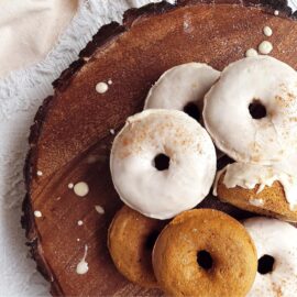 functional image pumpkin donuts top down on a dark slice of circular wood with bark around the edges. some donuts are glazed with coffee icing and come are plain donuts.