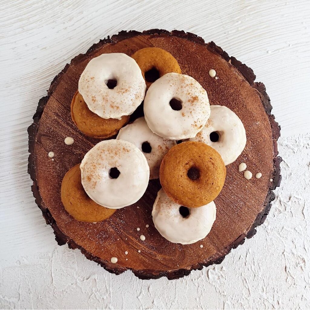 functional image glazed pumpkin donuts on a wooden circle with bark around the edges. background is white abstract. baked donuts are stacked on top of each other some have glaze and some do not for contrast