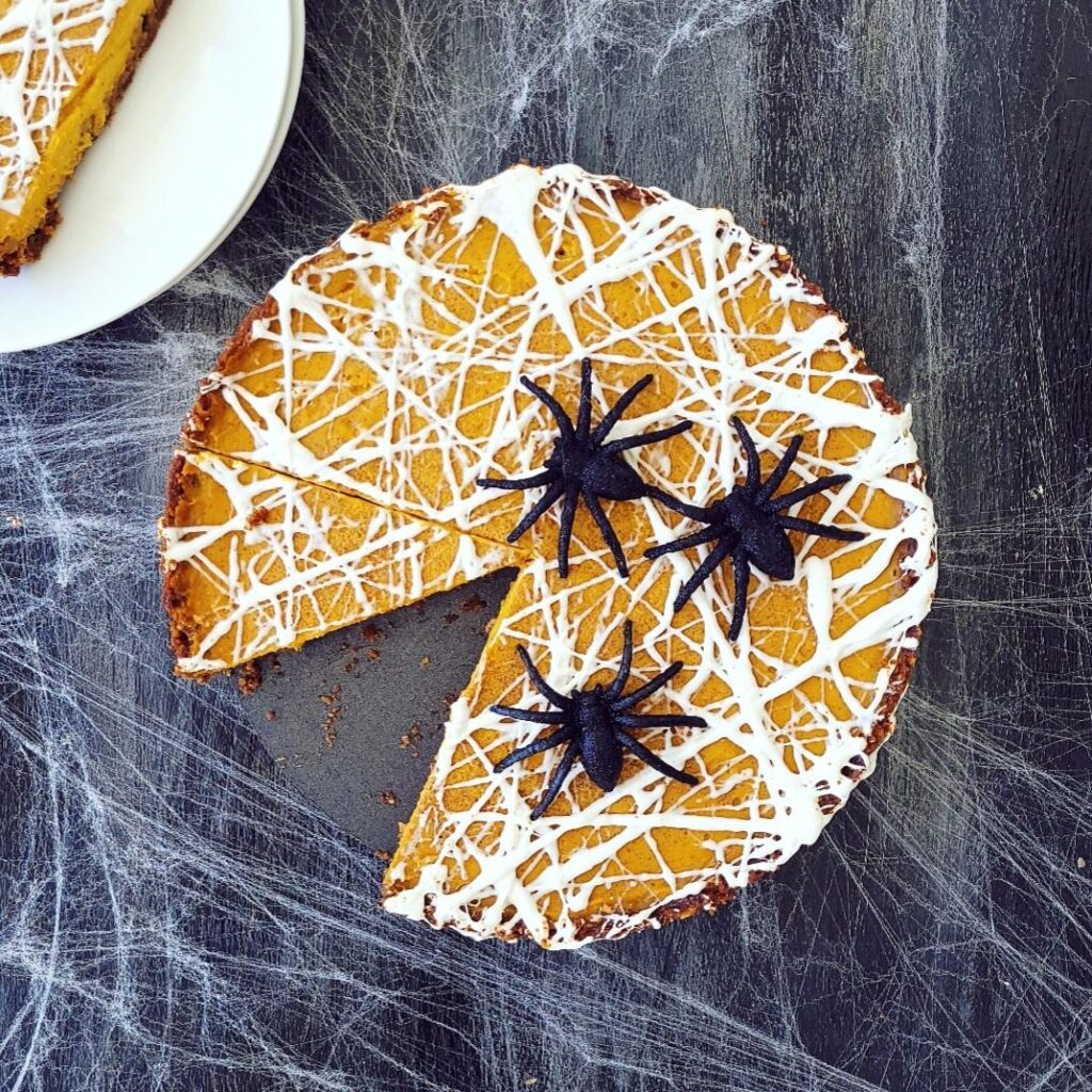functional image marshmallow spiderweb pumpkin tart halloween dessert top down on a black surface. one slice is cut from the tart and there are 3 black spiders on top
