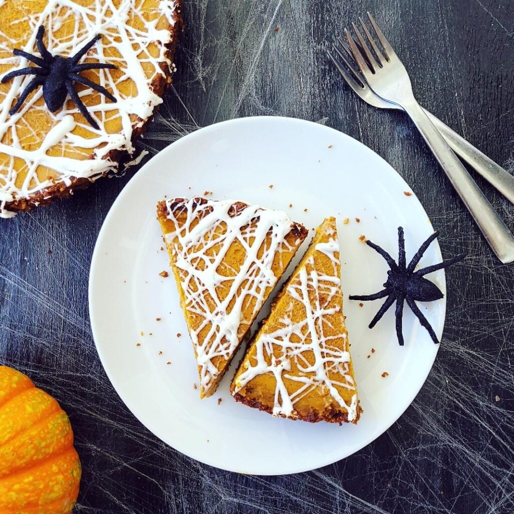 functional image pumpkin tart topped with a white marshmallow spiderweb  two slices cut on a shared white plate with a big black spider. top down background is black image is styled with a mini pumpkin two forks and the uncut portion of the tart in the upper left corner