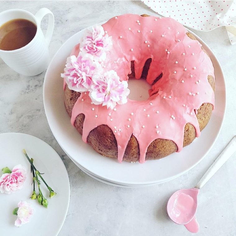 white chocolate raspberry bundt cake with pink glaze and pink carnations cake is uncut on a large white plate and has a cup of coffee next to it
