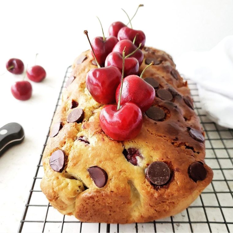 cherry chocolate chip bread loaf with giant chocolate chips and fresh cherries with stems garnishing the loaf on top. loaf is uncut and on a black baking rack