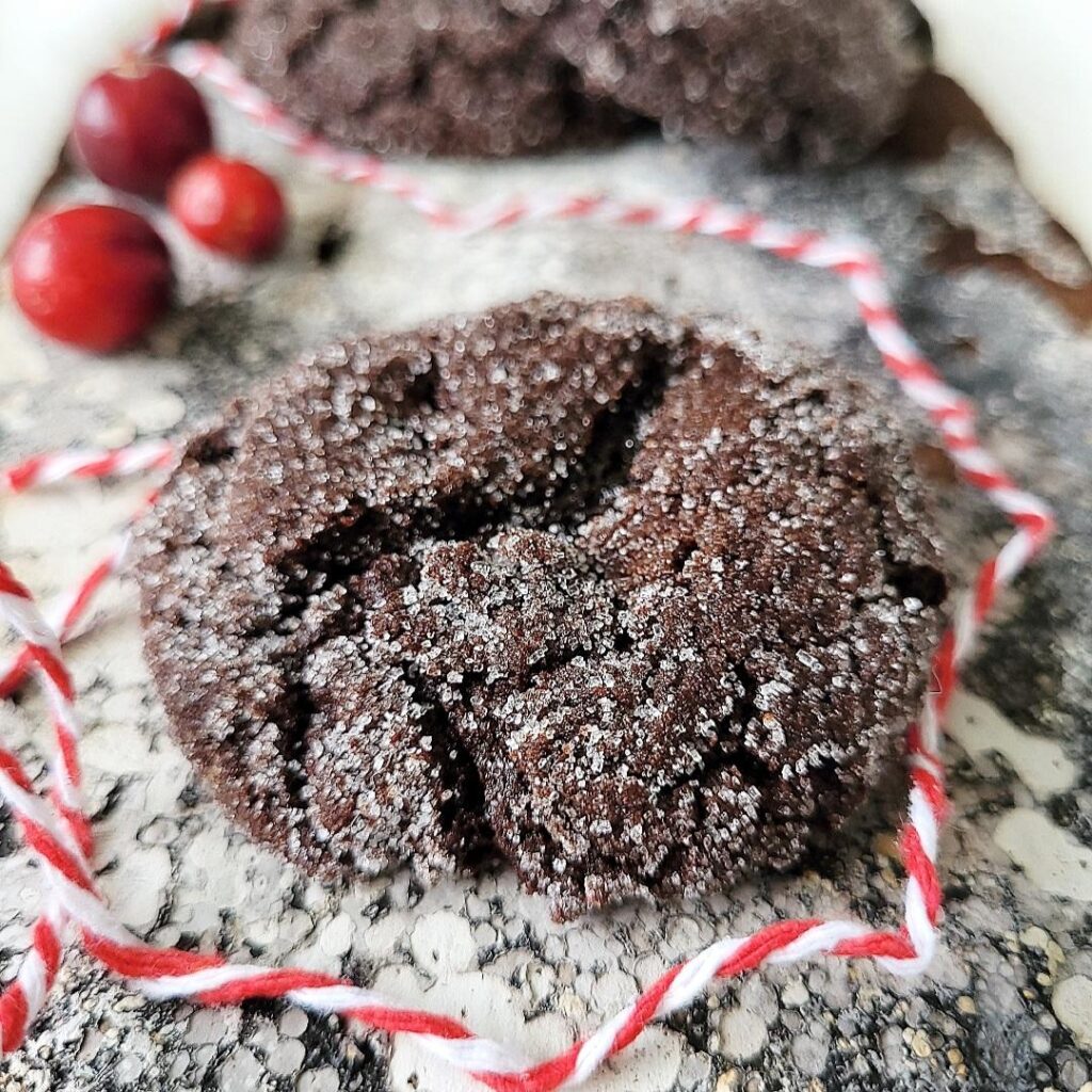 functional image chocolate molasses cookies sprinkled with sugar front, close up view of one cookie with two more in the background styled with cranberries and red and white holiday twine