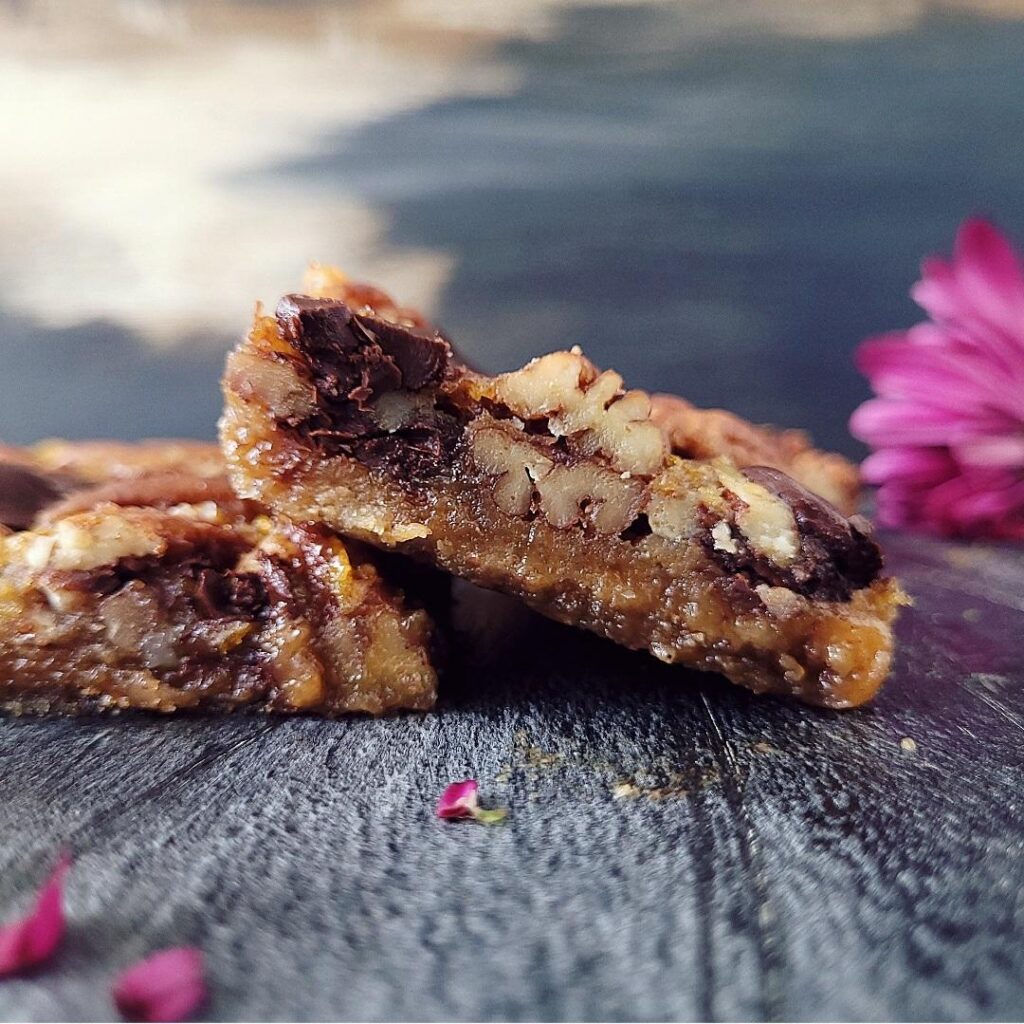 functional image chocolate chip pecan pie cookie bars side view of two bars cut into squares the left bar is sitting diagonally on the left bar there is a purple mum flower in the back right corner and the backdrop is blueish gray abstract