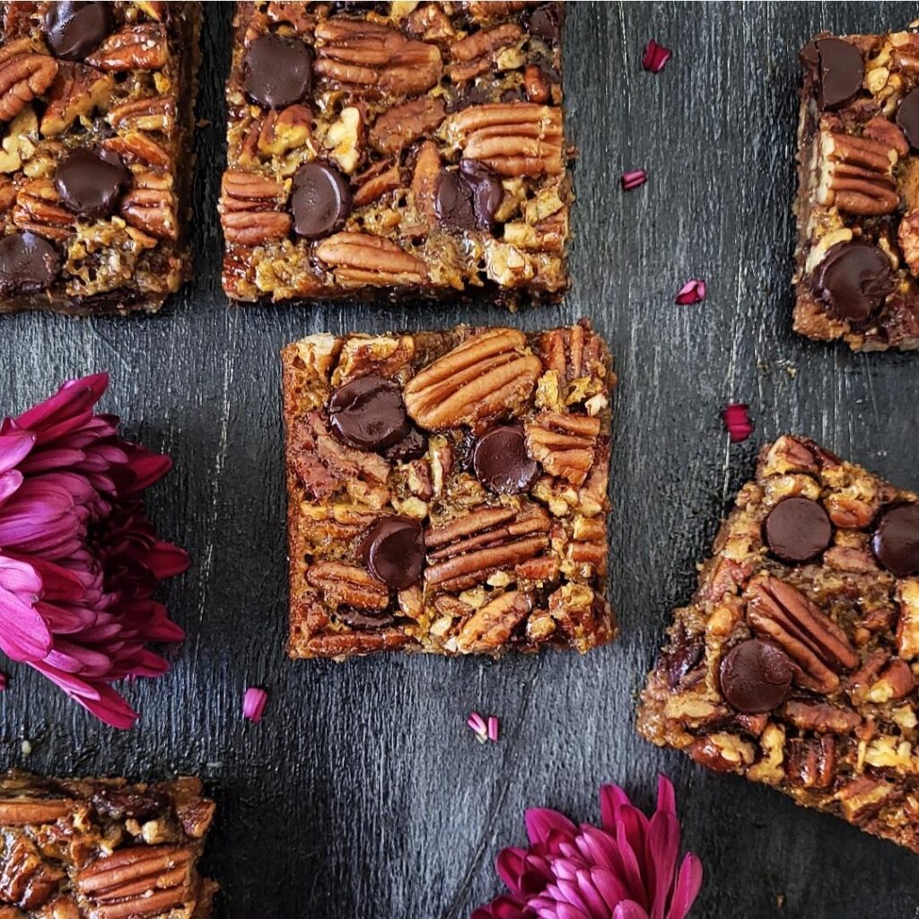 functional image chocolate pecan pie bars top down image on distressed black wood with purple mums. 6 bars cut into squares and scattered on the backdrop.