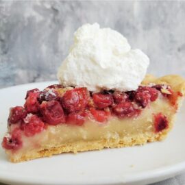 functional image cranberry custard pie one slice on a white plate with a big pile of whipped cream on top side view to see the crust, custard and cranberry layers background is wispy gray