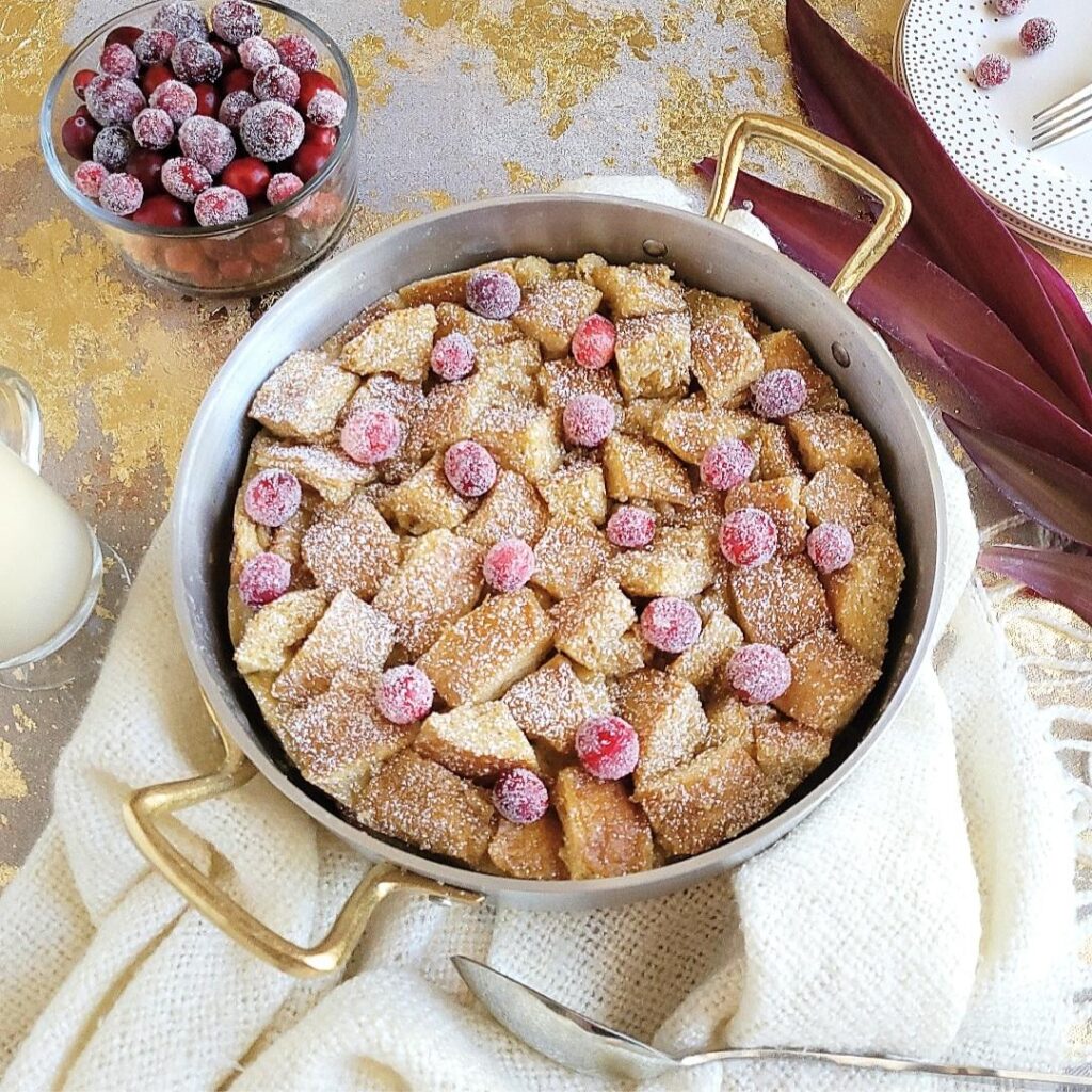 eggnog bread pudding garnished with fresh cranberries top down photo bread pudding is in a round silver baking pan with gold handles there is a bowl of sugared cranberries and gold polka dot plates