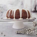 functional image eggnog bundt cake with white eggnog glaze on a white cake stand side view background is distressed gray and surface is decorated with sparkling silver baubles for christmas