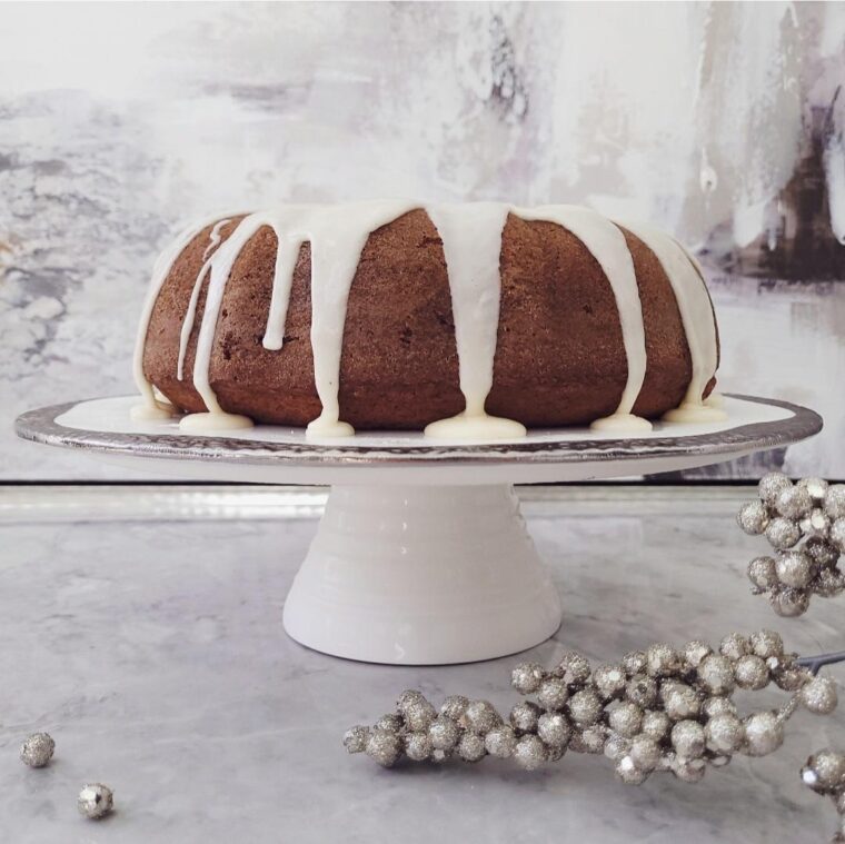 functional image eggnog bundt cake with white eggnog glaze on a white cake stand side view background is distressed gray and surface is decorated with sparkling silver baubles for christmas