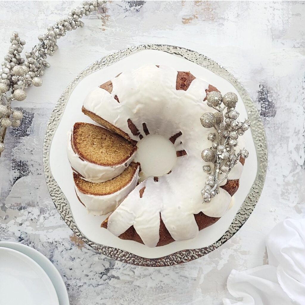 spiked eggnog bundt cake withe eggnog glaze top down on a sliver rimmed cake plate two slices are cut and askew cake is garnished with small silver baubles 