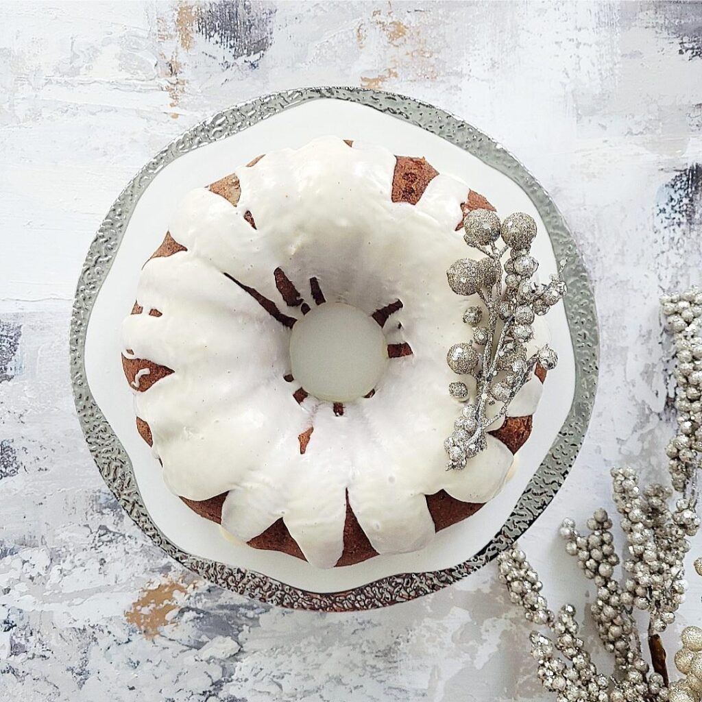 eggnog cake top down on a silver rimmed cake stand cake is topped with a white eggnog drip glaze and decorated with silver baubles for christmas