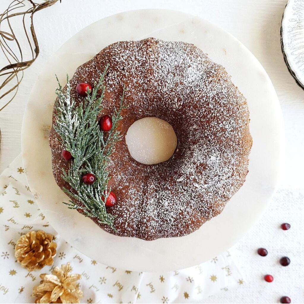 functional image gingerbread bundt cake top down photo. cake is garnished with snowy white powdered sugar evergreen twigs from a christmas tree and red cranberries. background is styled with gold pinecones and a white marble cake plate
