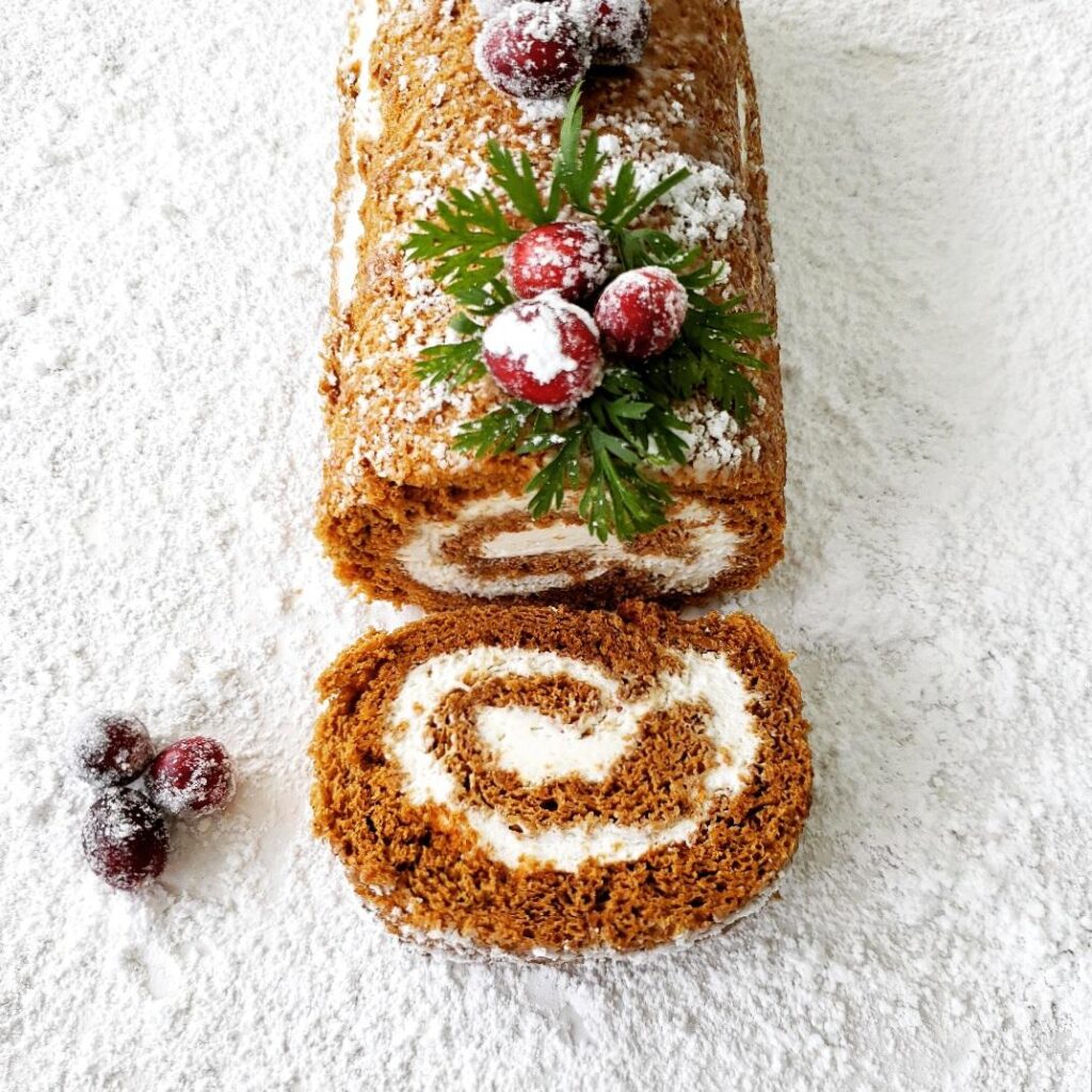 gingerbread cake roll 3/4 view with one slice cut so you can see the swirl of white eggnog filling cake is topped with cranberries, greenery and snowy powdered sugar on a white snowy surface