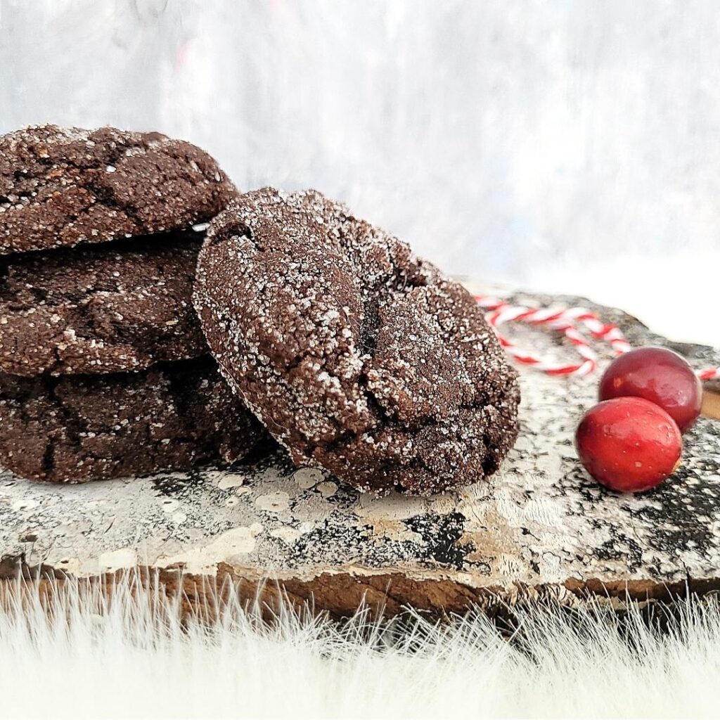functional image stack of three chocolate molasses crinkle cookies with a fourth cookie leaning on the stack. side view on a distressed wood surface styled with cranberries and red and white holiday twine