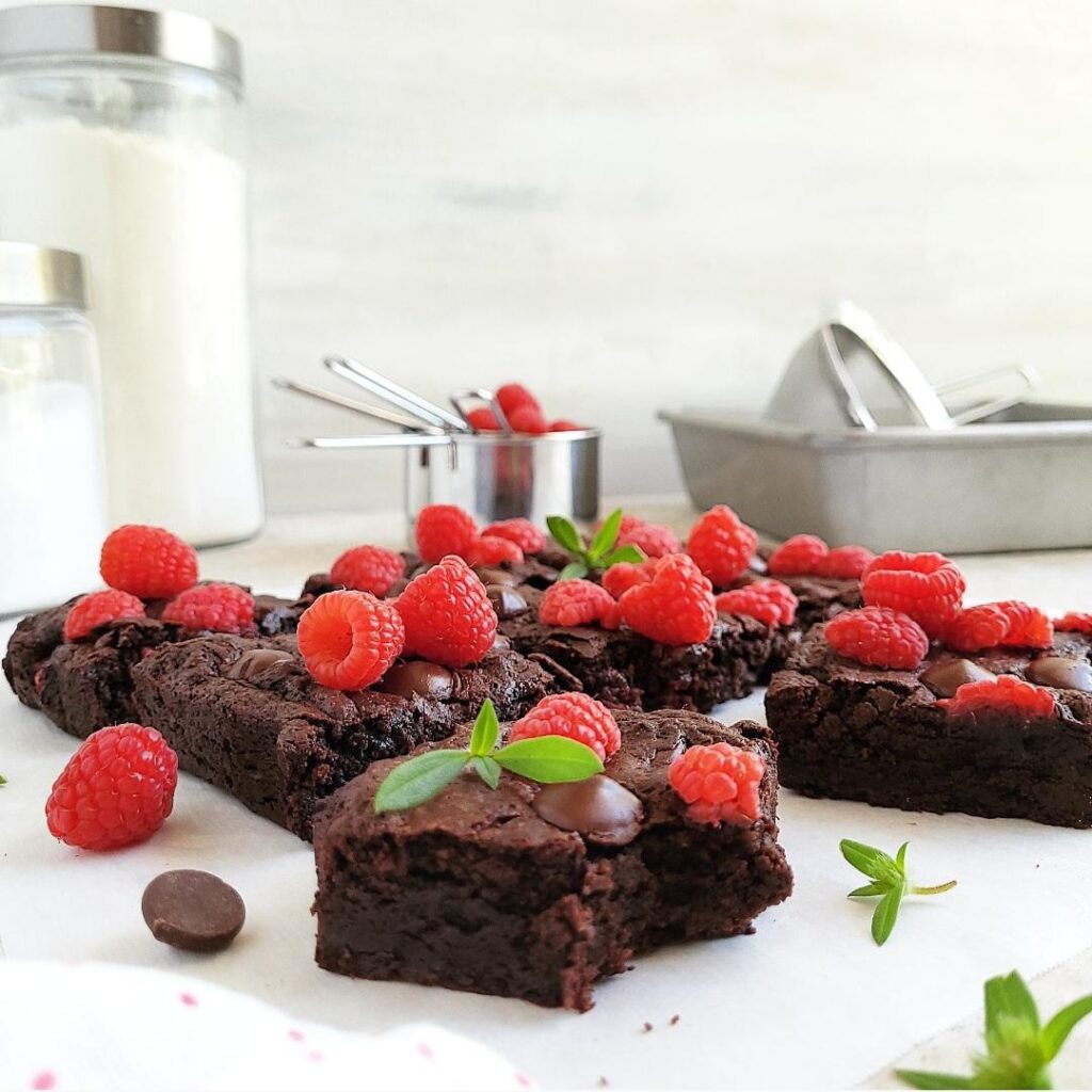 raspberry brownies cut into squares with fresh raspberries and giant chocolate chips on top background is gray with sugar and flour canisters, stainless steel measuring cups and aluminum baking pans. side view 