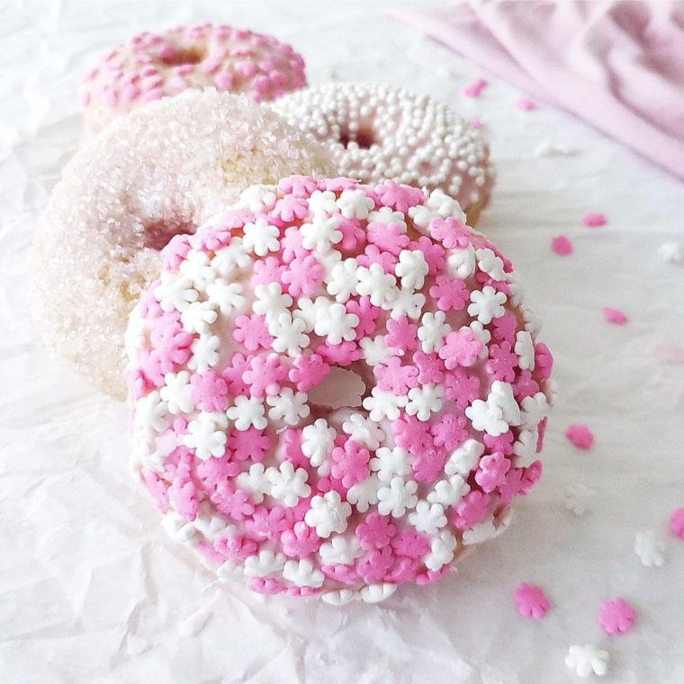 4 sugar cookie donuts each decorated with different toppings from pink and white snowflake sprinkles to clear sugar, white beads and pink hearts