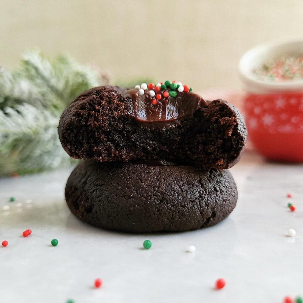 dark chocolate thumbprint cookies stacked two high the top cookie has a bite missing so you can see the interior crumb and ganache filling and holiday sprinkles background is christmas greenery and red and white bowls of sprinkles