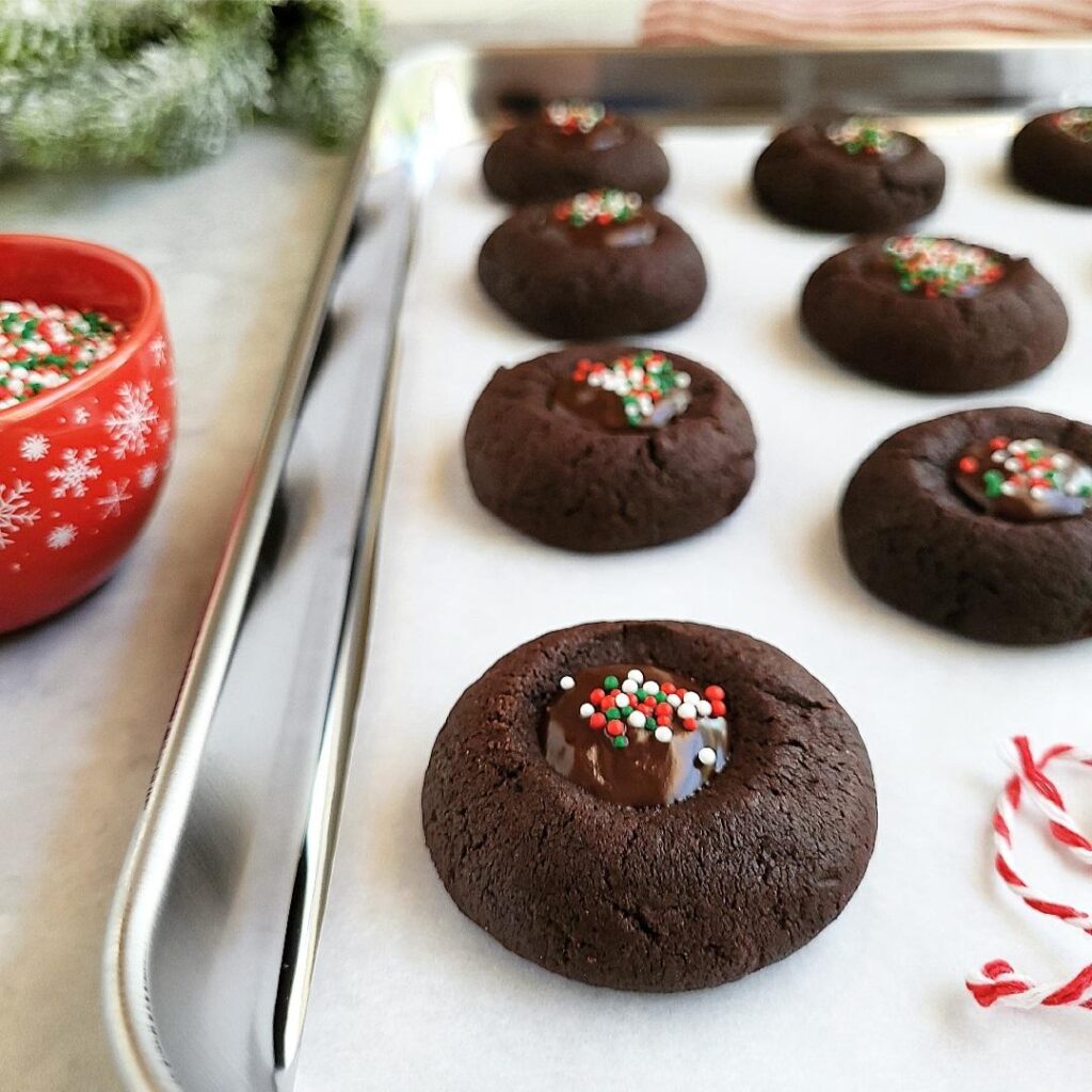 chocolate thumbprint cookies with chocolate ganache and christmas sprinkles lined up on a shiny baking sheet next to a red bowl of sprinkles and holiday greenery in the background