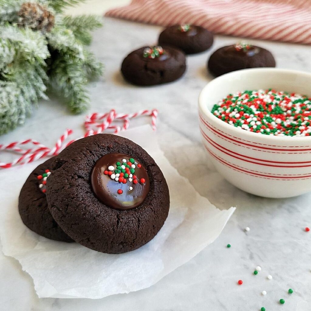 dark chocolate thumbprint cookies two cookies in forefront one is resting on the other so you can see the chocolate ganache and holiday sprinkles three more cookies are in the background between holiday greenery and a red and white striped linen there is a white bowl with red stripes filled with sprinkles 
