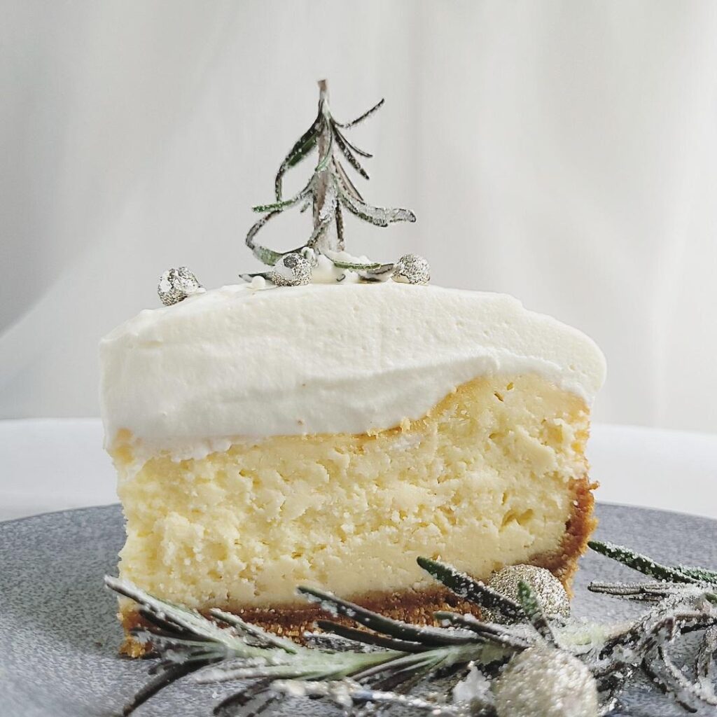 white chocolate christmas cheesecake with white chocolate whipped cream one slice on a gray plate side view with sugared rosemary "trees" and silver bauble decorations