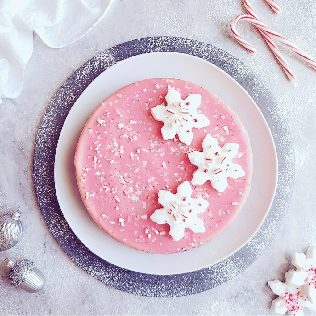 peppermint cheesecake top down photo of a pink ganache cheesecake with white snowflake marshmallows on top garnished with crushed candy canes this christmas cheesecake is aon a white plate on a gray charger and is styled with candy canes and silver ornaments