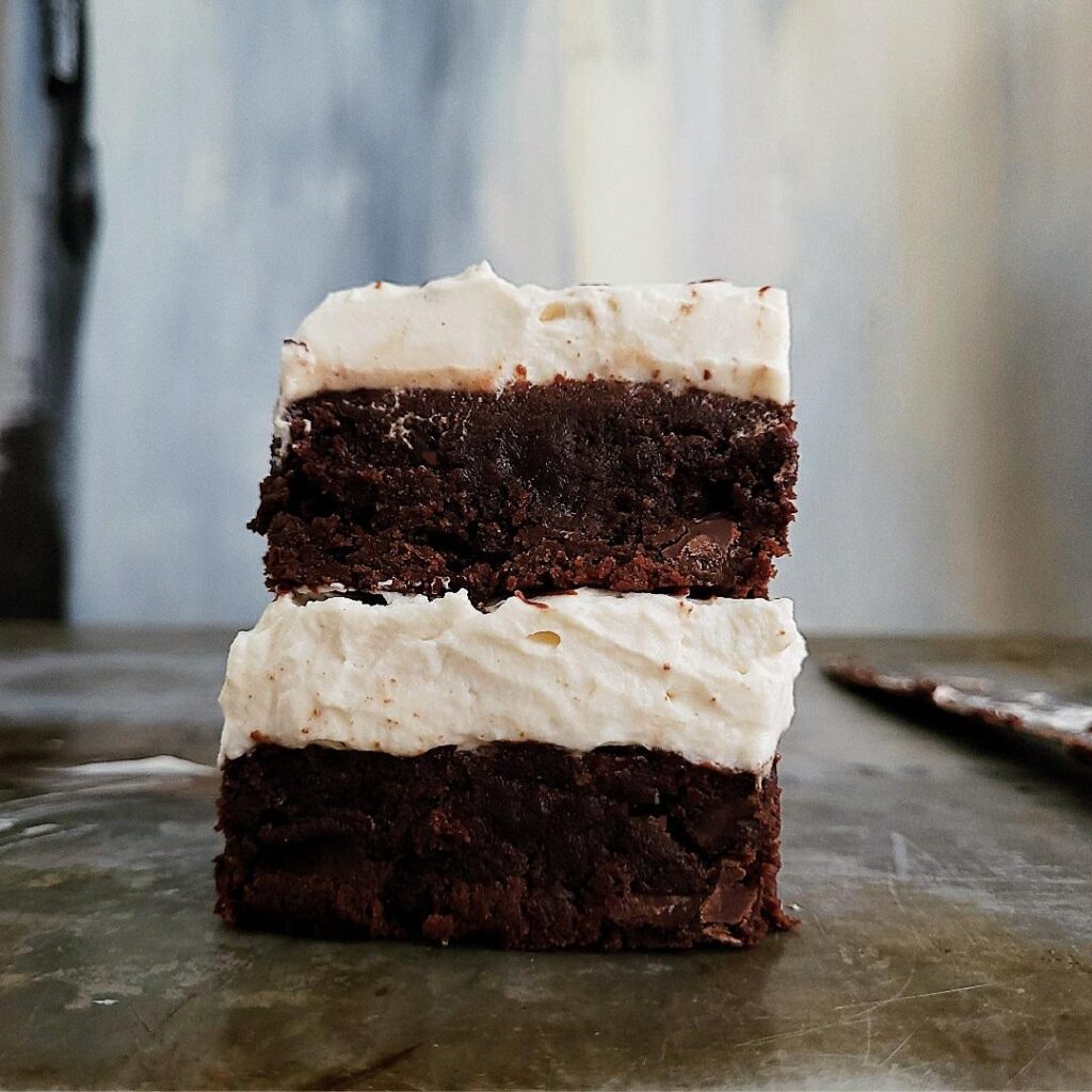 baileys brownies with baileys whipped cream stacked two high side view with blue abstract background on a weathered metal surface styled with a dirty knife