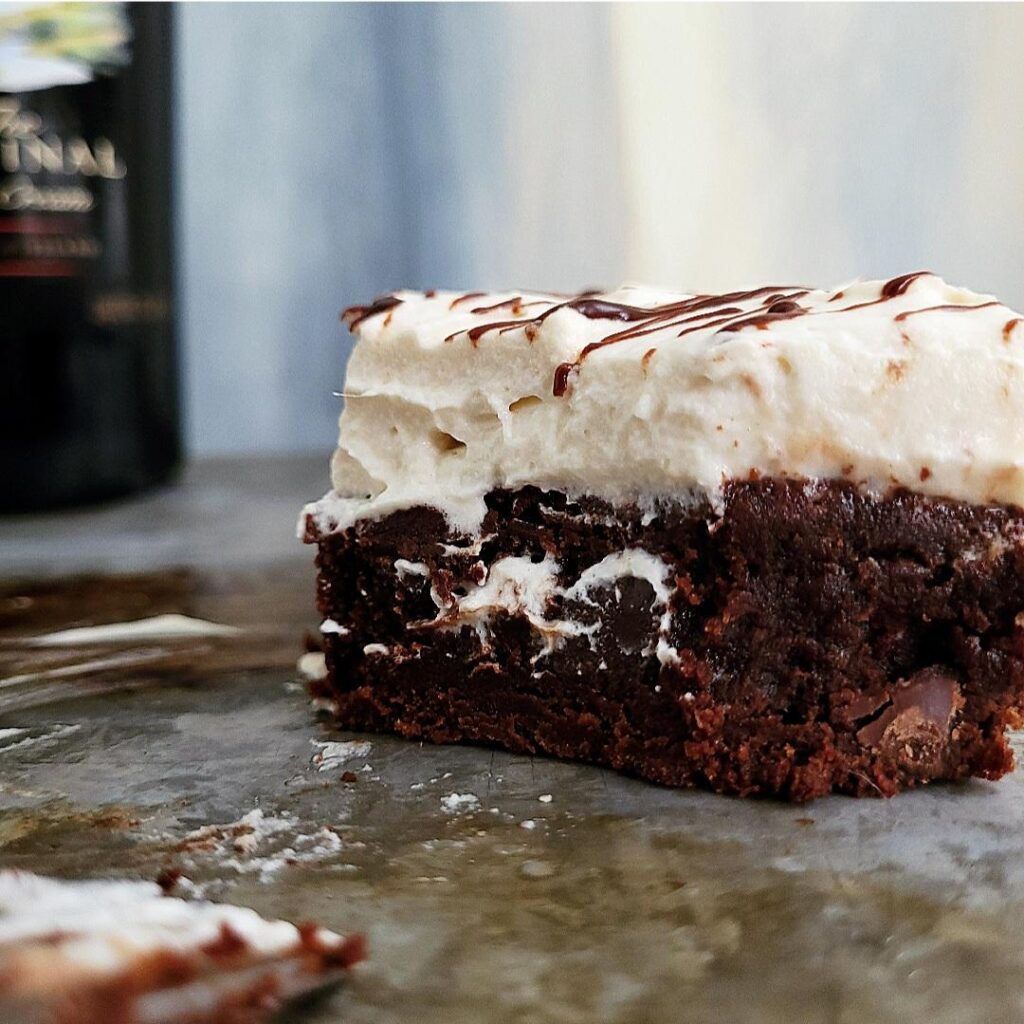 baileys brownies side view of a single brownie with baileys whipped cream on top. whipped cream is messy and has dripped down onto the side of the brownies. background is blue abstract. there is a bottle of baileys in the back left corner