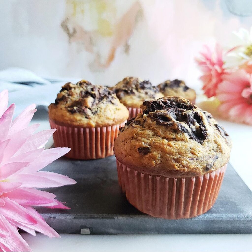 banana chocolate muffins on a gray slate board with pink cupcake liners and pink flowers close up view of 4 muffins