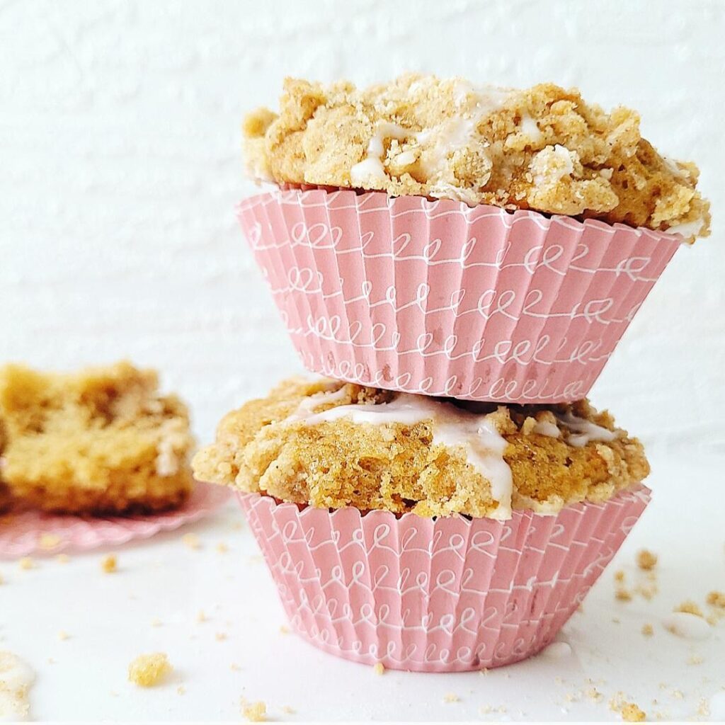 two coffee cake muffins with pink cupcake liners stacked 2 high. muffins have crumble topping and vanilla glaze. surface and background are white