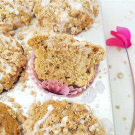 coffee cake muffins with crumble topping and white vanilla glaze in a shiny muffin tin one muffin is cut in half and propped up on a pink muffin liner so you can see the inner crumb there is a bright pink flower with a stem on the right side of the frame