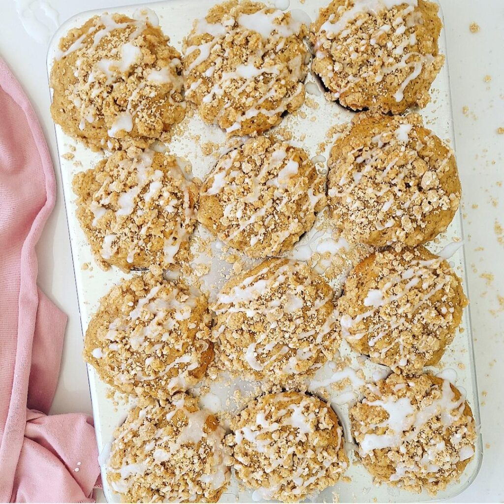 coffee cake muffins with crumble topping and vanilla glaze top down photo of 12 muffins in a shiny metal muffin tin surface is white and there is a pink linen on the left side of the screen 