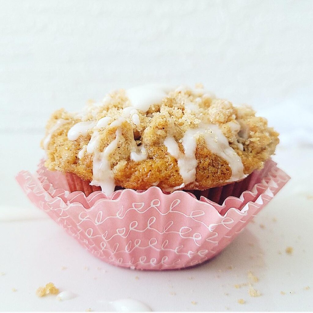 coffee cake muffin single muffin side view close up with a crumble topping and white venilla glaze muffin is wrapped in three pale pink muffin liners with white squiggly writing on them surface and background are white