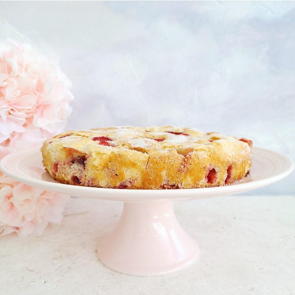 french strawberry cake uncut on a pale pink cake plate. side view there are pink flowers stacked on the left side of the frame. background is a wispy light blue and white abstract. surface is white limestone