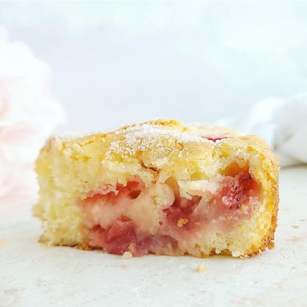 french strawberry cake side view of a single slice. close up view to see the delicate crumb, strawberries baked in and white sugar sprinkled on top background is pale blue and white