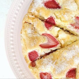 french strawberry cake top down view of cake on a pale pink cakestand cake is cut into three triangular slices so you can see the crackly top with fresh strawberries baked into the batter and the white sugar topping on this delicate cake