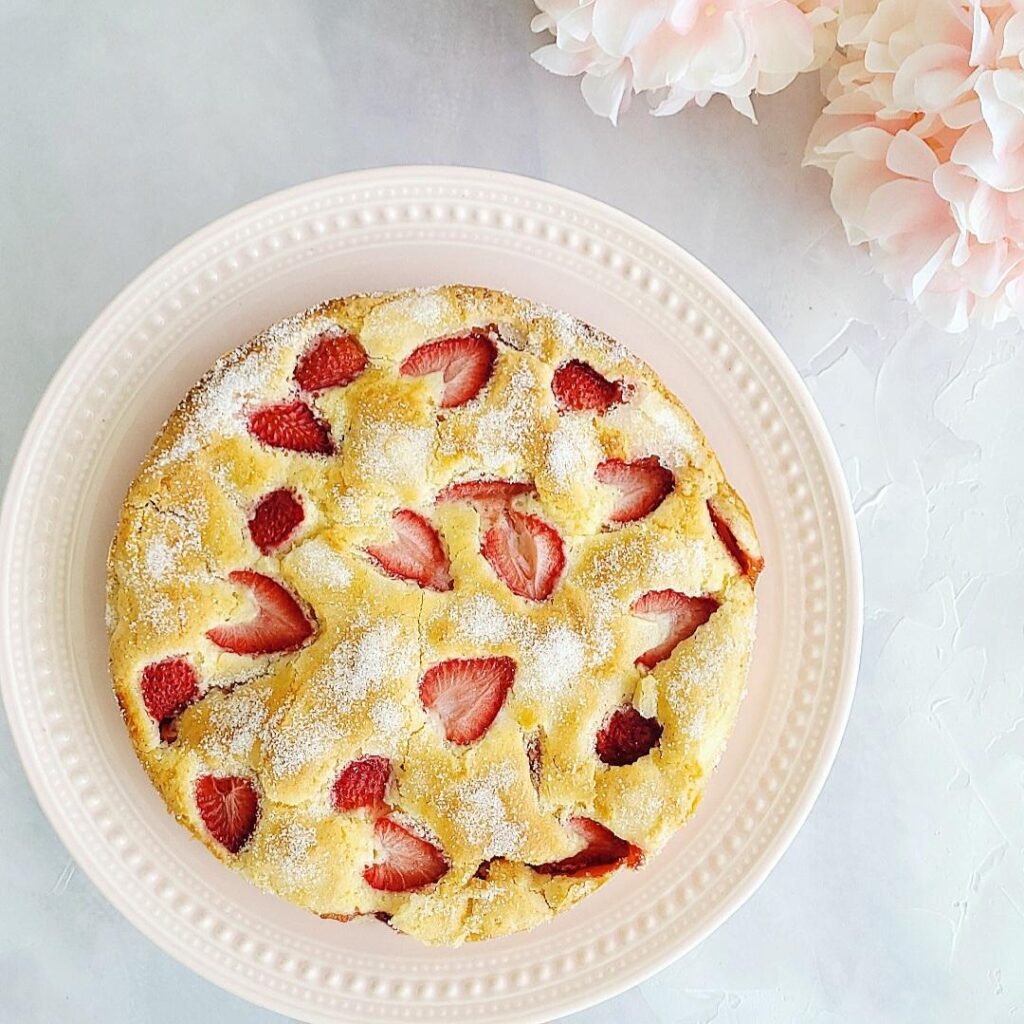 french strawberry cake top down view of cake on a pink cake plate. cake is spinkled with granulated sugar and features fresh strawberries peeking out of the baked cake background is light blue and white abstract and there are pink flowers in the top right corner