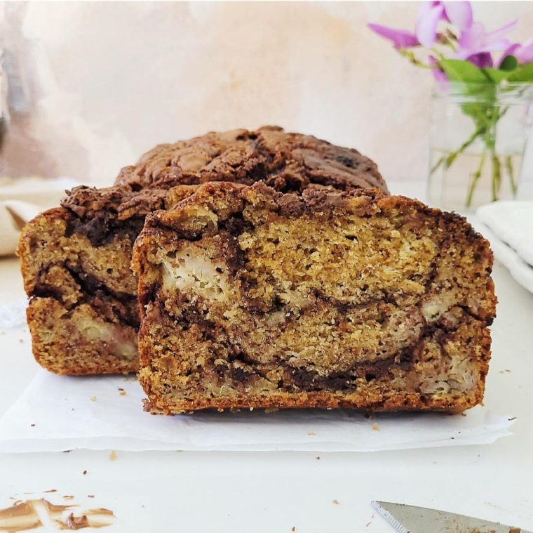 nutella banana bread side view of cut loaf so you can see the swirls of nutella, chunks of banana and moist crumb. background had fresh cut purple flowers in a clear class jar. 