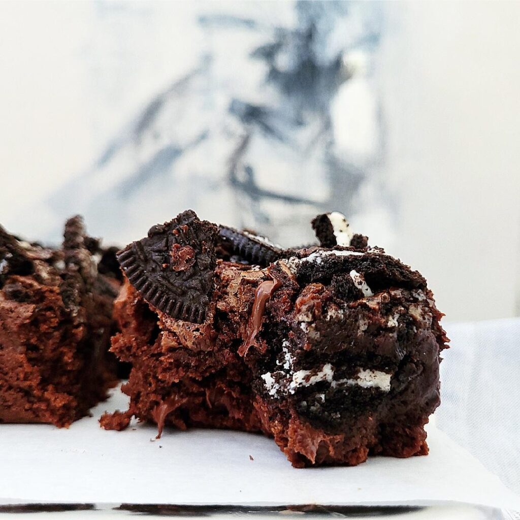 oreo brownies side view of a single square with a bite taken so you can see the fudgy center, melted chocolate chips and oreo cookies throughout and on top background is an abstract painting on canvas in gray black and white