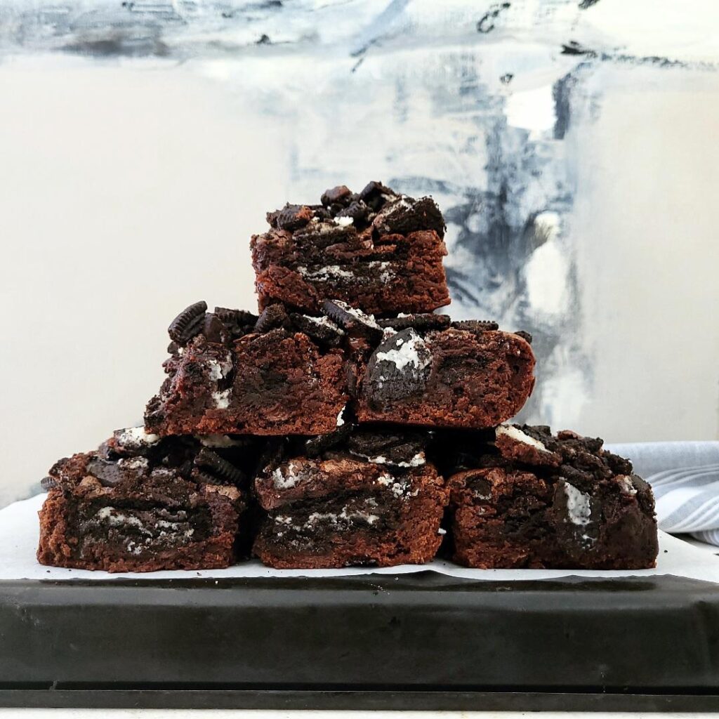 oreo brownies cut into squares and stacked in the shape of a pyramid three on the bottom, two in the middle and one on top background is abstract gray black and white canvas painting