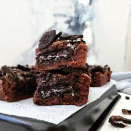 oreo brownies side view of two brownie squares stacked on top of each other so see the layers of brownie, cookies and chocolate chips stack in on a black baking pan covered with white parchment background is an abstract canvas painting with gray, white and black