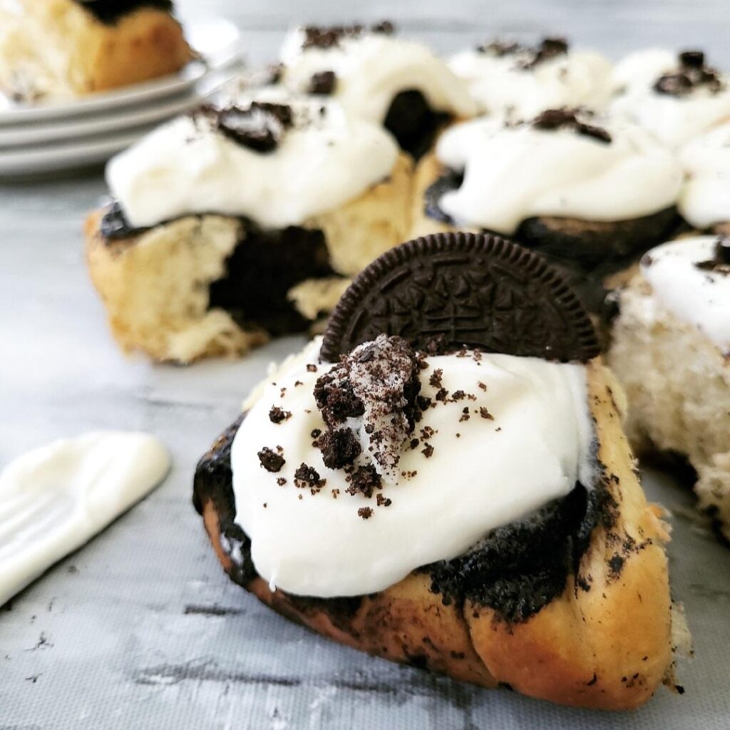 oreo cinnamon rolls full frame of cookies and cream cinnamon rolls one is in the background on a stack of plates. all others are topped with cream cheese icing and oreo cookie crumbs. one is  featured in the front with half an oreo cookie propped on top.